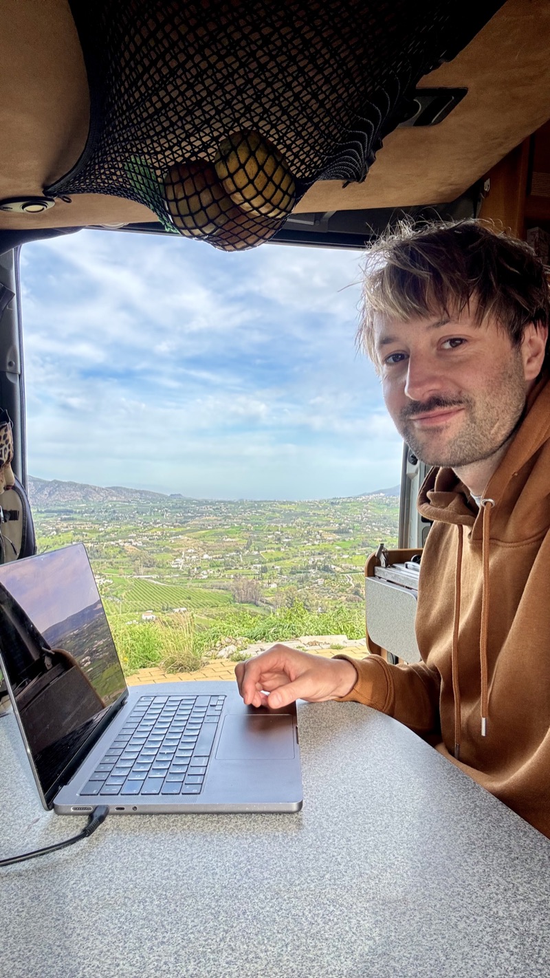 Jascha working on a laptop in his camper van, the open door framing the Andalusian valley below